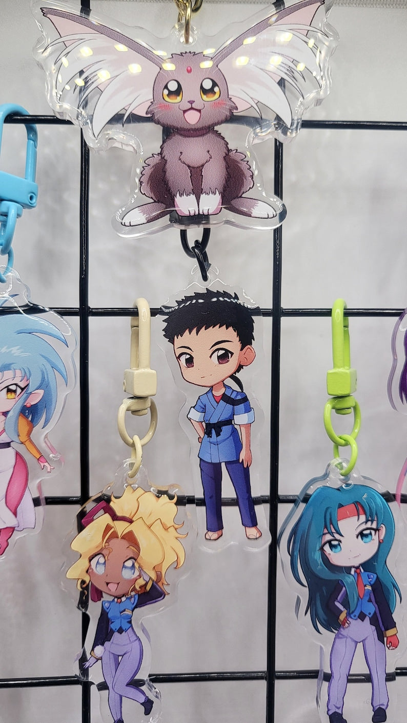 Exclusive Tenchi Muyo acrylic keychain featuring double-sided designs of Ryo-Ohki, Tenchi, Sasami, Ryoko Hakubi, Washu, Ayeka, Mihoshi, and Kiyone. Vibrant, scratch-resistant colors with a glitter epoxy coating for a standout look. Perfect for accessorizing backpacks, lanyards, and Ita bags. Durable silver D-clasp for long-lasting use. A must-have collectible and a perfect gift for Tenchi Muyo and anime fans!